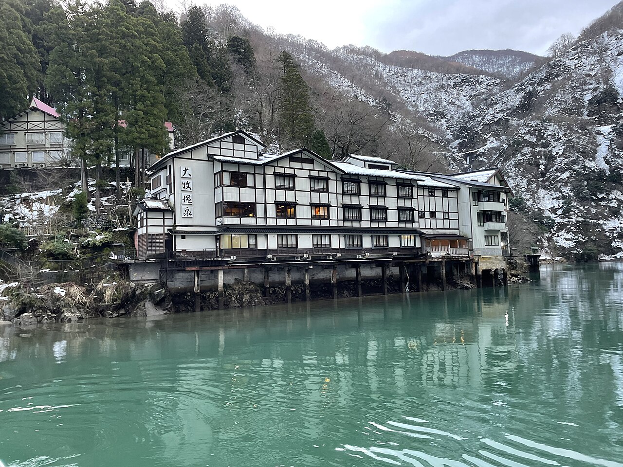 雪の残る山を背に、大牧温泉の白い建物が青緑の水面に映る